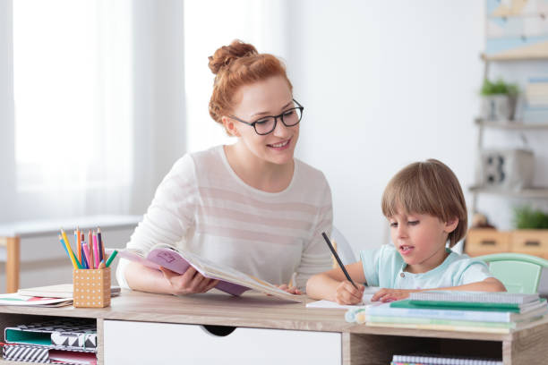 Smiling mother helping son with exercises of math while sitting at desk with notebooks