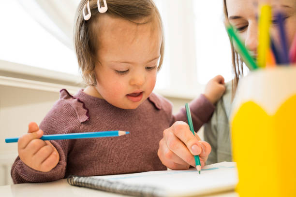 Cute girl with down syndrome drawing and sitting over a blank sheet of paper at the table. Girl spending time with her mother and sharing positive emotions with her. Stock photo